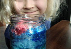 Uśmiechnięta dziewczynka i wykonany przez nią „kosmos” w szklanym słoiku z przewagą niebieskiego brokatu